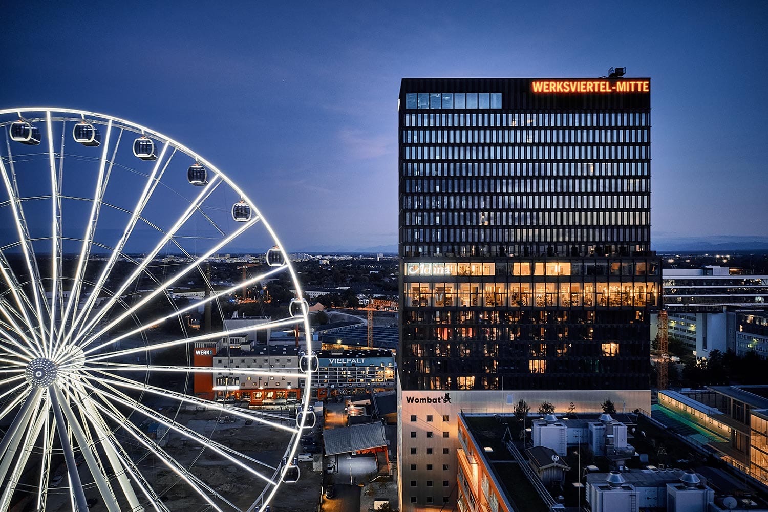 Adina Apartment Hotel Munich lit up at dusk, the city ferris wheel beside it
