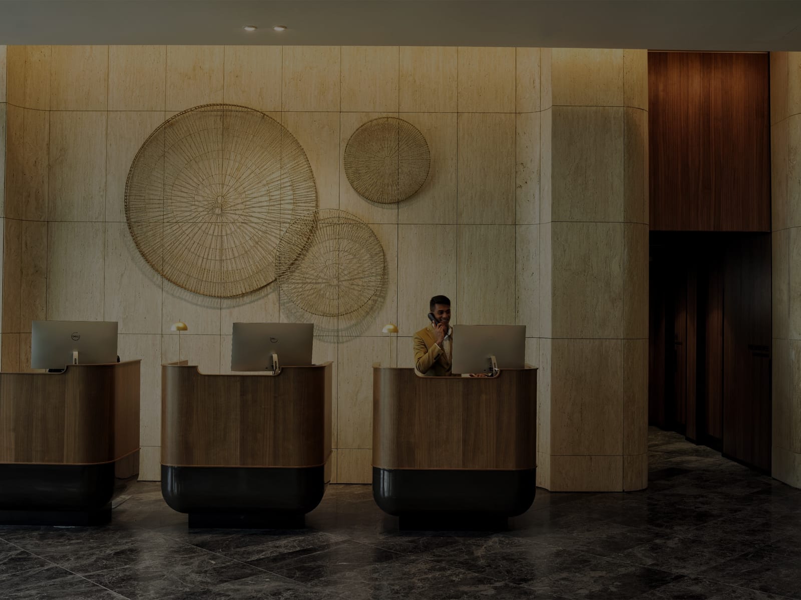 A TFE Hotels lobby: three timber check-in pods against a stone wall hung with woven discs, a team member taking a call at the far pod.